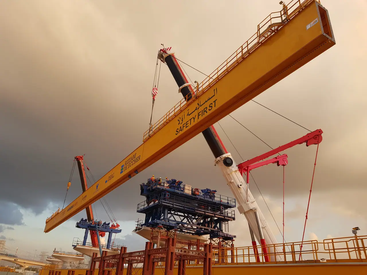 Two mobile cranes lifting a yellow bridge-launching girder marked SAFETY FIRST under a dusk sky