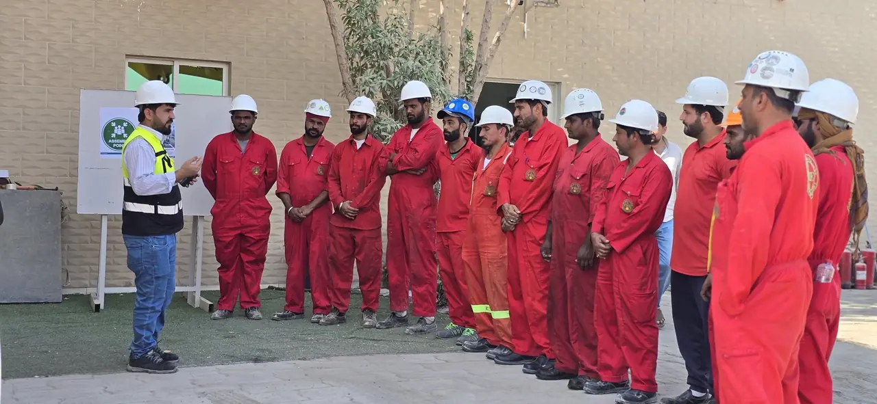 An officer briefing a line of FHS crew in red coveralls beside the emergency assembly point sign