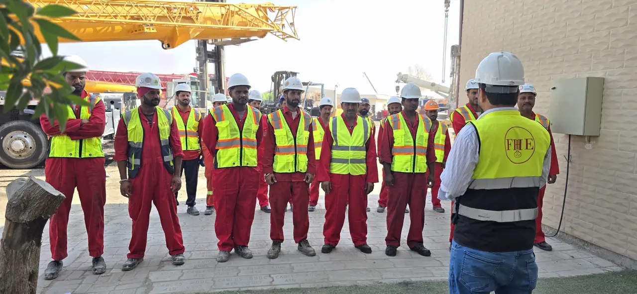 An officer in an FHE vest, seen from behind, addressing the crew, with a yellow crane boom beyond