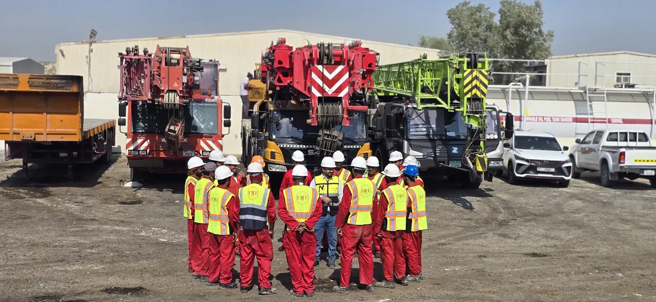 FHS crew in red coveralls and FHE hi-vis vests gathered in a circle for a toolbox talk in front of three mobile cranes