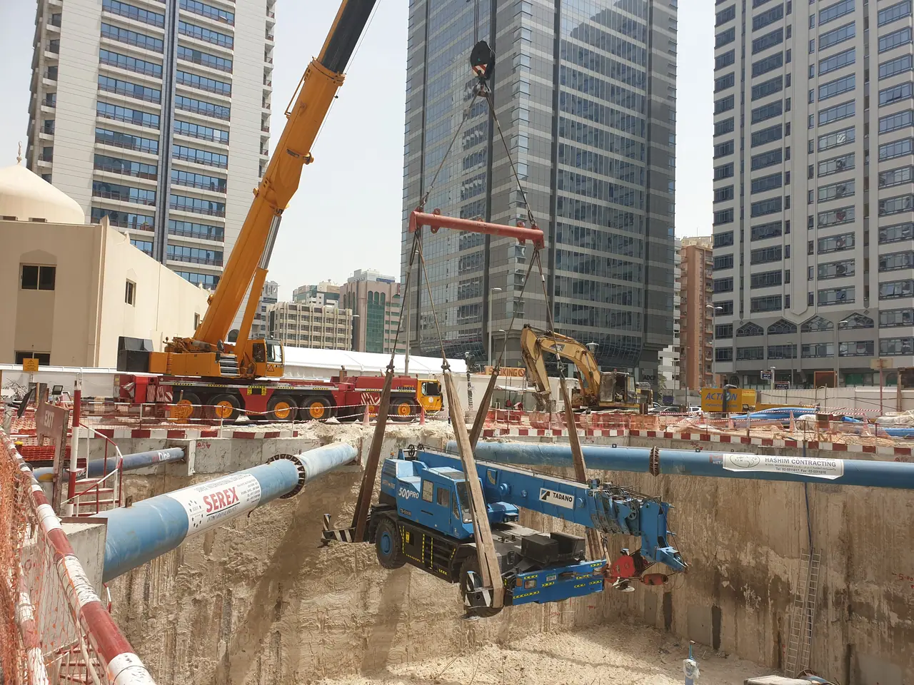 Mobile crane lowering a piling rig into a deep excavation between city towers